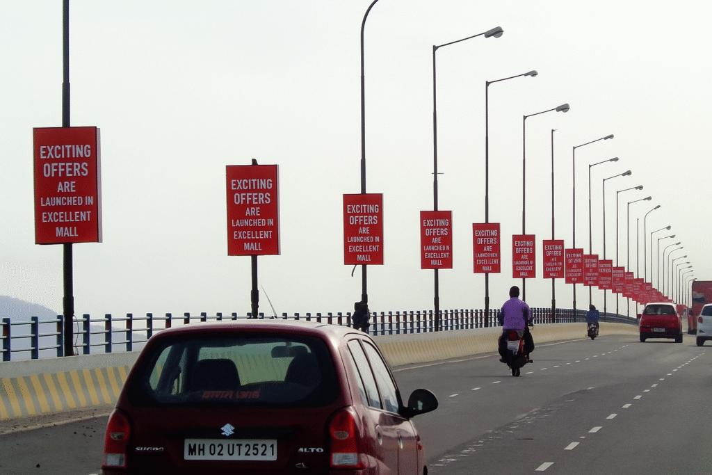 Red pole kiosk banners on lamp posts promoting “Exciting Offers at Excellent Mall,” with vehicles and a red Suzuki Alto on a city road.
