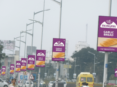 A row of digital pole kiosk advertising companies in Lucknow displaying advertisements for 'Monginis Garlic Bread,' showcasing images of garlic bread slices, set along a busy highway.
