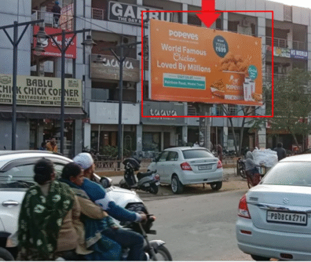 Street view near PPR Mall Model Town with 20x10 ft Popeyes hoarding by ACME Advertising Co., promoting Ludhiana launch at PVR Flamez Mall; visible shops, parked vehicles, motorcycles, and pedestrians